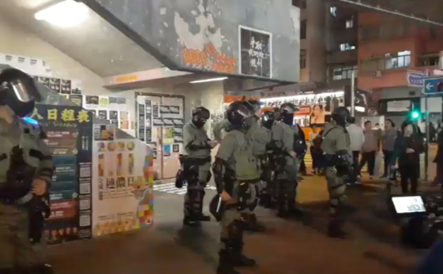 Polizisten in Schutzausrüstung stehen vor einer Menge von Helmträgern mit Waffen, mit Plakaten an Wänden, Deckenleuchten, Gebäuden und einem Verkehrszeichen mit Text im Hintergrund, in Hong Kong während einer Protestauflösung.