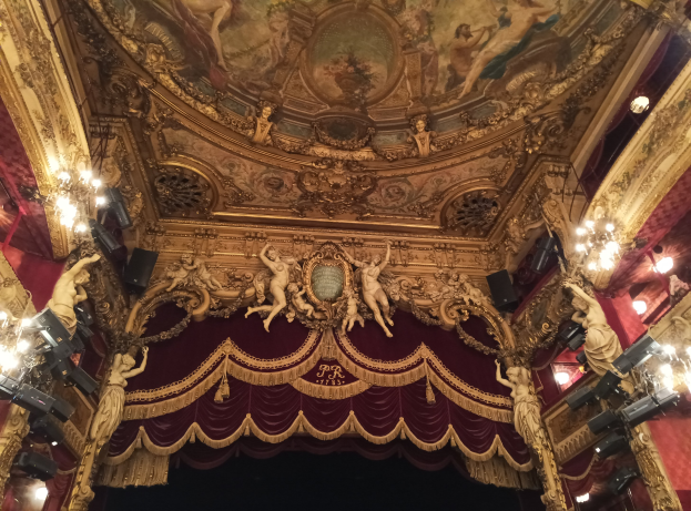 Theaterinnenraum mit roter Gardine, Kronleuchtern, Skulpturen, Lampen und einer bemalten Decke, mit einer Bühne und einigen Menschen unten.