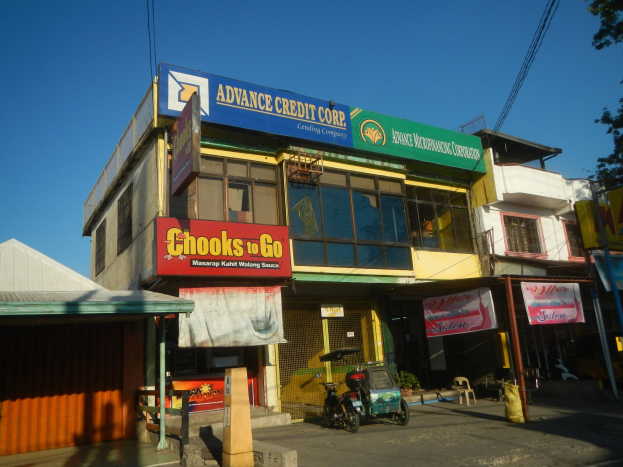 Ein Geschäftsgebäude mit einer Tafel, auf der "Advance Credit Corp" steht, das sich neben einer Straße mit Fahrzeugen, Werbetafeln, Strommasten, Kabeln, Bäumen und einem klaren blauen Himmel befindet.