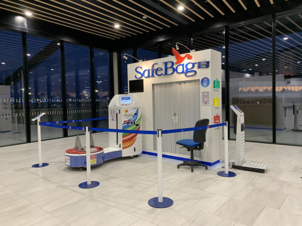 Sicherheitskontrolle für Gepäck am Flughafen mit einem Stuhl, Absperrgittern, Maschinen, einem Bildschirm, einer bedruckten Tafel, Glaswänden, Deckenlampen und sichtbarem Boden.