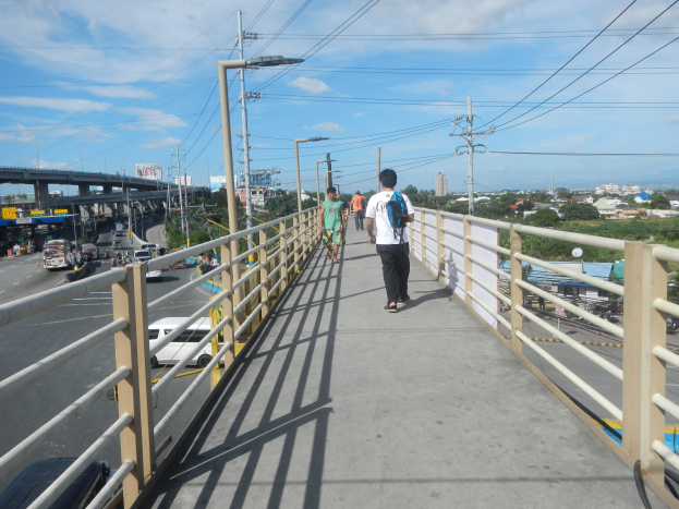 Eine Gruppe von Menschen überquert eine Fußgängerbrücke über eine vielbefahrene Straße, mit Geländern, Strommasten und Drähten im Hintergrund, während Fahrzeuge darunter fahren und Gebäude, Bäume und ein bewölkter Himmel zu sehen sind.