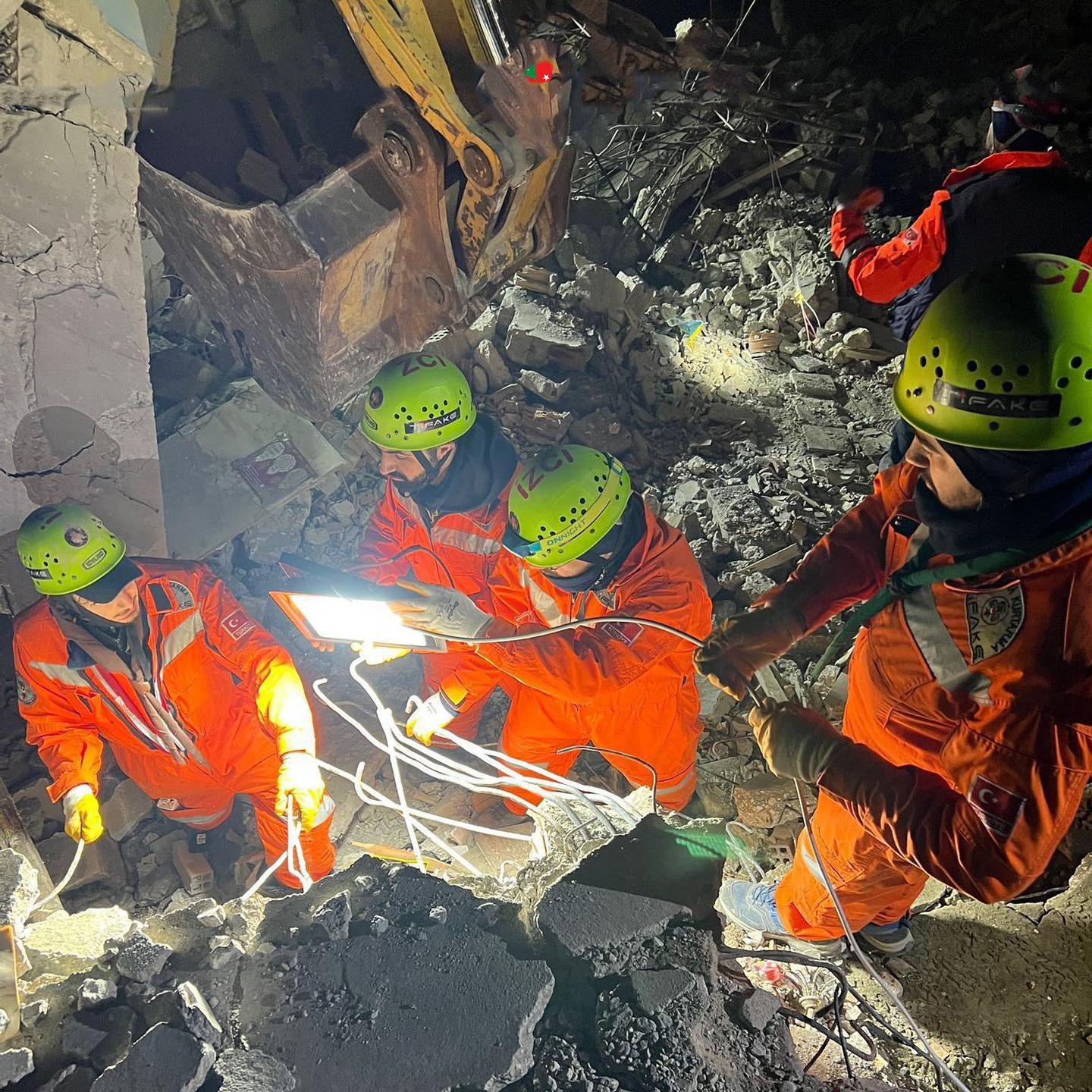 Gruppe von Menschen in orangefarbenen Uniformen und grünen Helmen bei der Arbeit an Schutt mit einem Kran im Hintergrund bei einem Einsatz nach dem Einsturz eines Gebäudes.