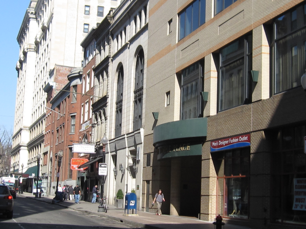 Eine belebte Stadtstraße mit hohen Gebäuden, Fahrzeugen, Fußgängern, einem Hydranten, Laternen, Bäumen und einem klaren blauen Himmel.