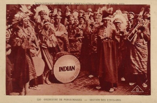 Schwarz-weißes Foto einer Gruppe von Menschen in Kostümen mit Musikinstrumenten, mit einer Trommel in der Mitte und Text und Logo unten.