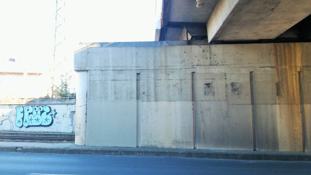 Betonwand mit Graffiti, eine Straße darunter, ein Gebäude und ein Turm im Hintergrund und ein klarer blauer Himmel.