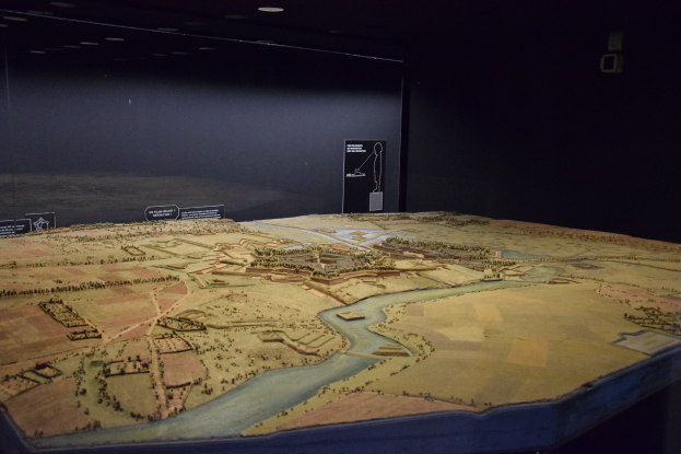 Ein Stadtmodell in einem Museum mit einem Tisch im Vordergrund und einer Texttafel im Hintergrund unter beleuchteter Decke.