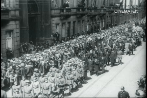 Schwarzes Foto einer Parade mit einer großen Menge Menschen, die eine Straße entlangmarschieren, einige halten Gewehre in den Händen, vor einem Gebäude mit einem sichtbaren Wasserzeichen.