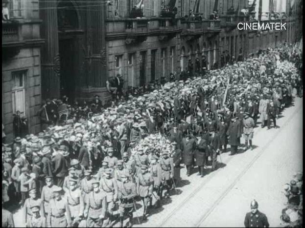 Schwarzes Foto einer Parade mit einer großen Menge Menschen, die eine Straße entlangmarschieren, einige halten Gewehre in den Händen, vor einem Gebäude mit einem sichtbaren Wasserzeichen.