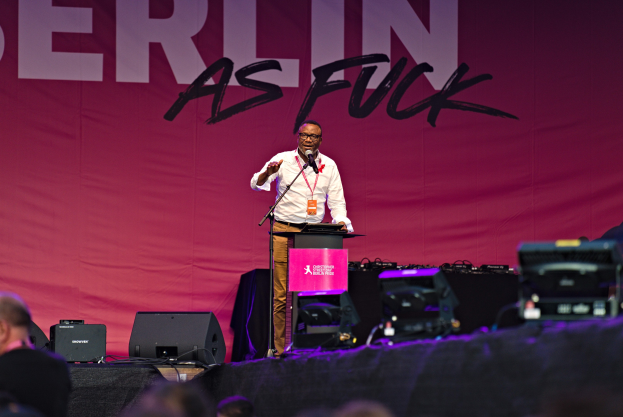 Ein Mann in weißem Hemd und braunen Hosen steht auf einer Bühne und hält ein Mikrofon, hinter ihm eine pinkfarbene Fahne mit der Aufschrift "Berlin as Fuck" und Lautsprecher auf der Bühne.