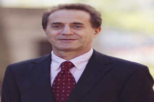 Ein selbstbewusster Mann im Anzug und mit Krawatte, der wie ein Generalstaatsanwalt aussieht, steht lächelnd vor einem Gebäude.