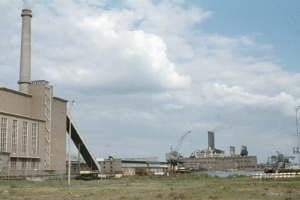 Ein altes Foto einer Fabrik mit Rauch, der aus ihren Schornsteinen aufsteigt, umgeben von Gebäuden, Kränen, Polen, Lampen und Schildern, mit grasbewachsenem Boden und einem bewölkten Himmel.