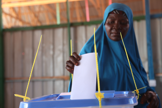 Frau in blauem Kopftuch, die ein Blatt Papier hält und vor einer Kiste mit herausragenden Stöcken steht, vor einer Wand und einem Schuppen an einer Wahllokal im Hintergrund.