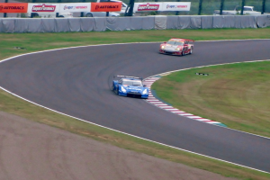 Zwei Autos auf einer Rennstrecke mit Gras auf beiden Seiten, Schilder, Absperrungen, Pfosten und Bäumen im Hintergrund.