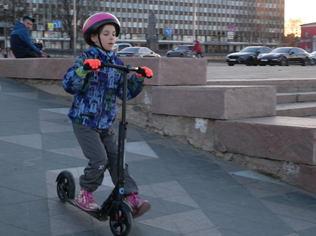 Junger Junge in Helm und Handschuhen auf einem Scooter auf einem Gehweg mit Treppen, Fahrzeugen, Menschen, Bäumen, Pfählen, Brettern, Gebäuden und einem klaren blauen Himmel im Hintergrund.