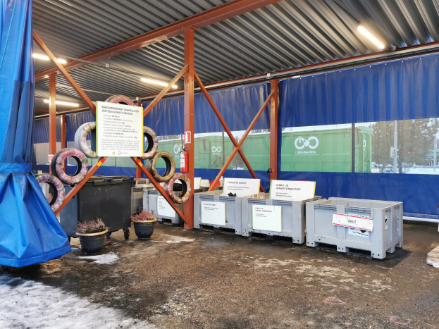 Großes Lagerhaus-Innere mit verschiedenen Reifen, Containern, Topfpflanzen, bedruckten Platten, Pfählen, Beleuchtung, einem blauen Tuch und anderen Gegenständen, gesetzt in einer schneebedeckten Outdoor-Umgebung mit Bäumen und Fahrzeugen.