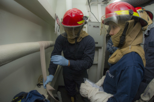 Drei Feuerwehrleute in Schutzausrüstung stehen zusammen in einem Raum mit Rohren, einer Tasche und Maschinen im Hintergrund.
