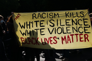 Eine Gruppe maskierter Personen hält ein Transparent mit der Aufschrift "Rassismus tötet Weiße Stille, weiße Gewalt, Schwarze Leben zählen" auf einer dunklen Straße.