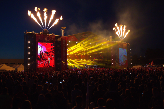 Eine große Menge bei einem Musikfestival schaut auf einer hell erleuchteten Bühne performende Künstler, mit Zelten links und Feuerwerk in einem dunklen Himmel dahinter.