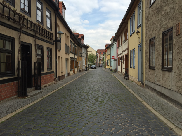 Eine Kopfsteinpflasterstraße in Heidelbergs Altstadt, gesäumt von Gebäuden mit Fenstern und Türen, mit parkenden Fahrrädern an den Seiten, Bäumen im Hintergrund und einem bewölkten Himmel.
