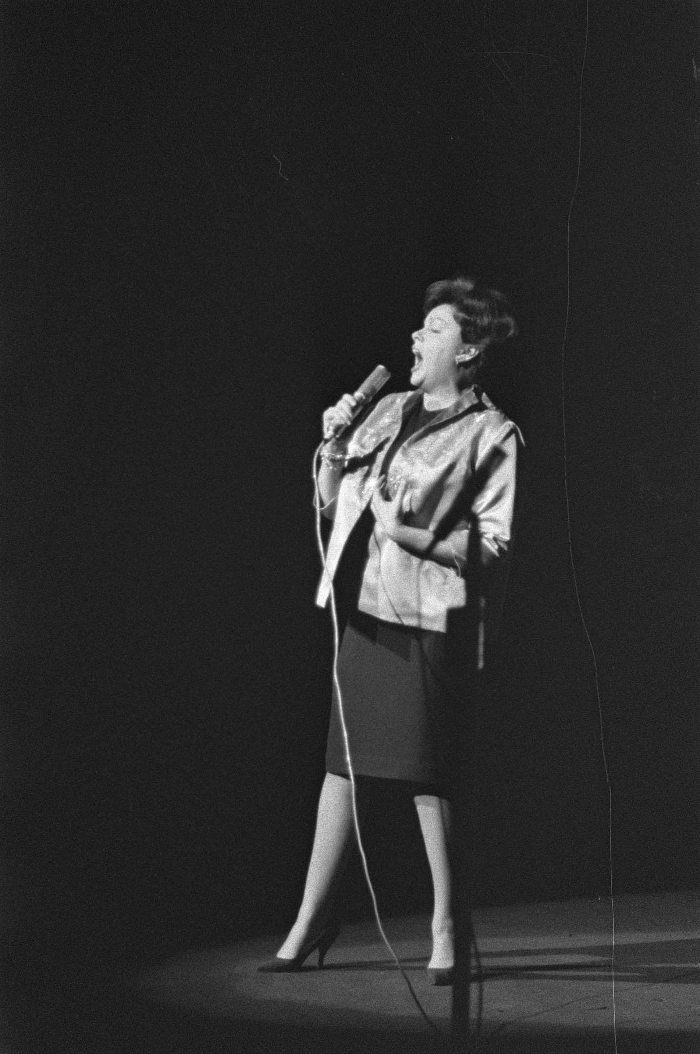 Eine Frau in einem schwarzen und weißen Outfit aus den 1950er Jahren singt fröhlich auf einer von einem Scheinwerfer beleuchteten Bühne in ein Mikrofon.