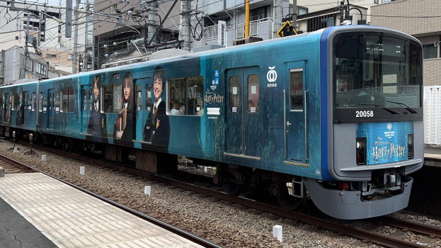 Blauer Zug fährt auf Schienen neben einem Bahnsteig mit Oberleitungen und Masten, Gebäude im Hintergrund unter einem klaren Himmel.