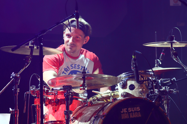 Ein Mann in einem roten und weißen T-Shirt spielt Schlagzeug auf der Bühne während eines Konzerts, grinsend umgeben von Mikrofonen.