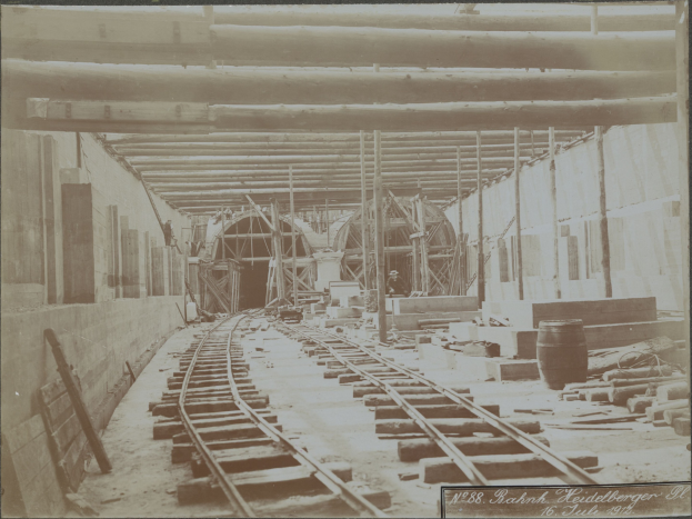 Schwarz-weiß-Foto einer Eisenbahnschiene in einem Gebäude unter Bau, mit verstreuten Holzgegenständen, Fässern, Wänden, Pfählen und anderem Schutt; Text unten sichtbar.