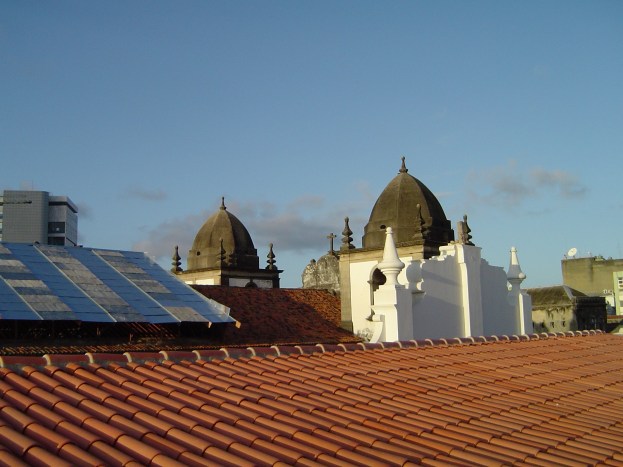 Stadtlandschaft mit Geb├Ąuden im Vordergrund, Solarpanels auf einem Dach und einem blauen Himmel im Hintergrund.