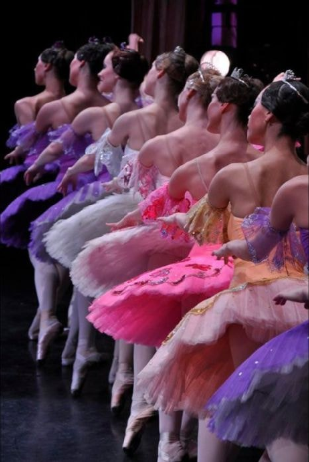 Balletttänzerinnen in rosafarbenen Tutus sitzen auf einer Bühne, beleuchtet von einem Licht im Hintergrund, bewegen sich in harmonischer Grazie.