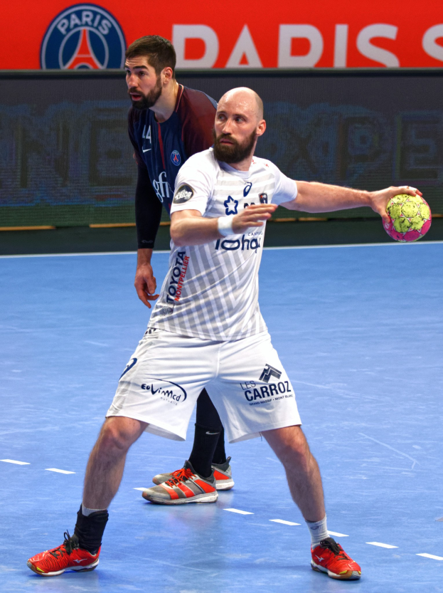 Zwei Männer spielen Handball auf einem Platz, einer hält den Ball, mit einem Hintergrundschild, auf dem "Paris Saint-Germain vs Paris Saint-Germain" steht.