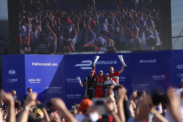 Gruppe von Menschen auf einem Formel-E-Podium in Mexiko-Stadt mit Pokale, Sponsor-Plakaten und einem Bildschirm im Hintergrund.
