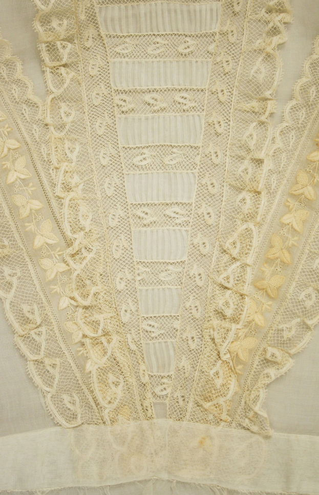 Nahaufnahme eines weißen Kleides mit zarter, detailreicher Spitzenverzierung in verschiedenen Formen und Mustern, aus leichtem, luftigem Sommergewebe.
