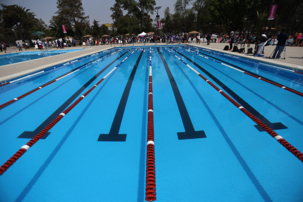 Großes Schwimmbad mit Bahnentrennung, umgeben von Menschen mit Taschen und Gegenständen, mit Zelten, Bannern, Stangen, einem Schild und einem bewölkten Himmel im Hintergrund.