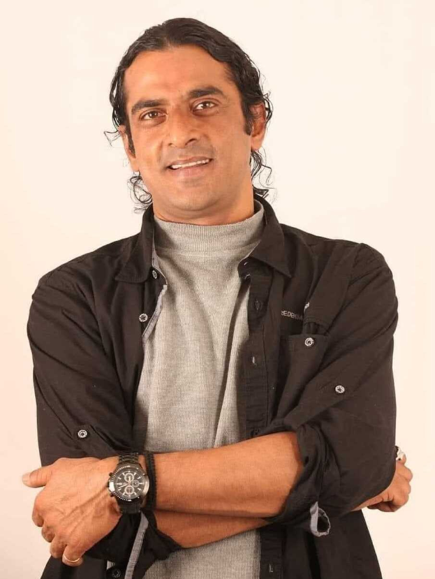 Ein Mann in einem schwarzen und aschfarbenen Outfit mit überkreuzten Armen, der eine Uhr trägt und vor einem weißen Hintergrund steht, in Gedanken versunken.