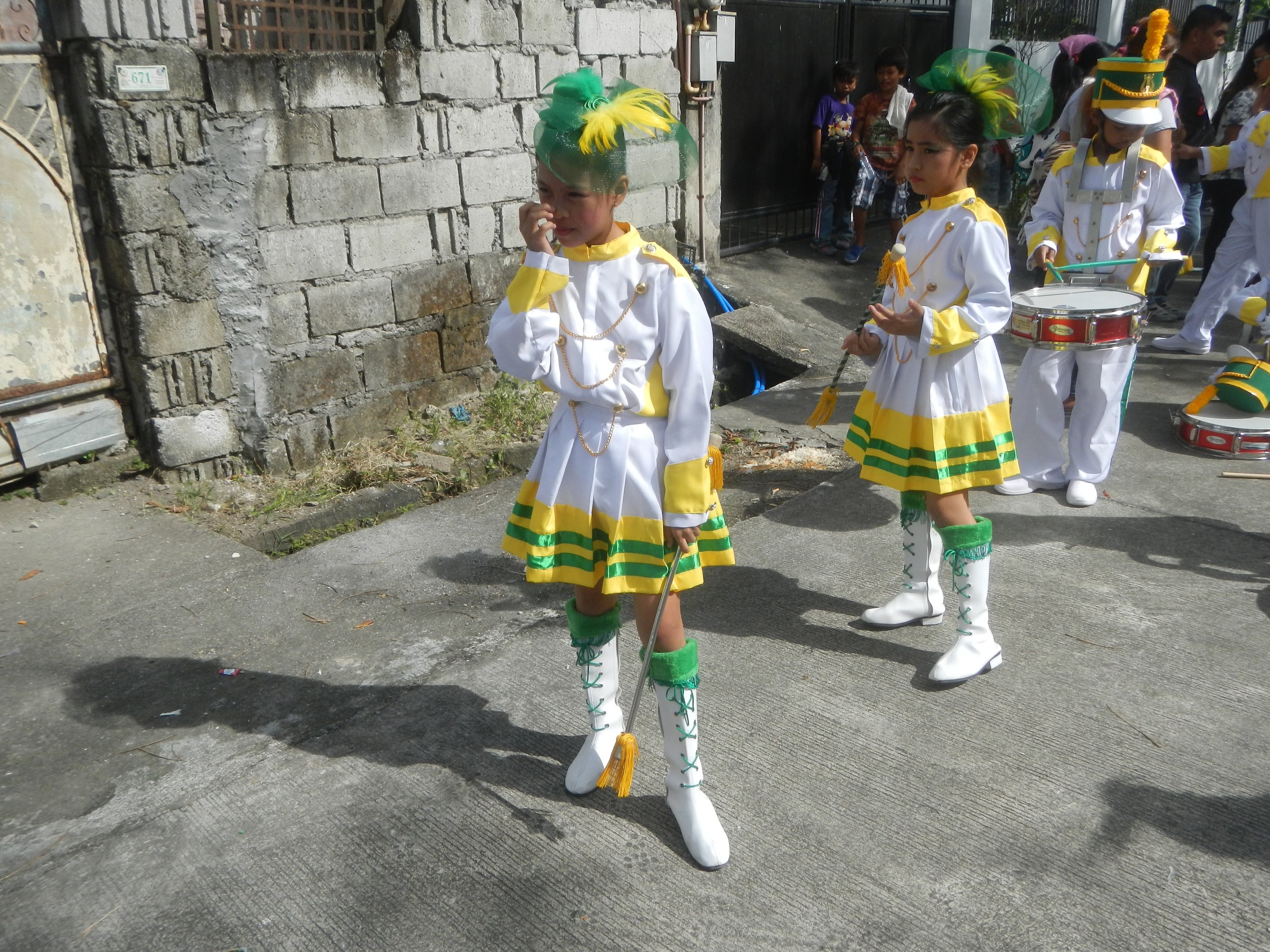 Gruppe junger Mädchen in weißen und gelben Outfits marschiert, während sie Musikinstrumente spielen und an einer Wand, einem Tor und Bäumen vorbeikommen.