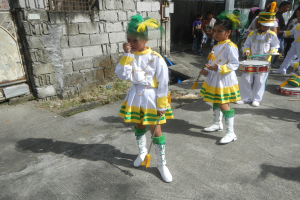 Gruppe junger Mädchen in weißen und gelben Outfits marschiert, während sie Musikinstrumente spielen und an einer Wand, einem Tor und Bäumen vorbeikommen.