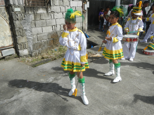 Gruppe junger Mädchen in weißen und gelben Outfits marschiert, während sie Musikinstrumente spielen und an einer Wand, einem Tor und Bäumen vorbeikommen.