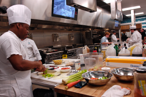 Eine Gruppe von Köchen in einer Küche beim Zubereiten von Speisen auf einem Schneidbrett, wobei ein Koch im Vordergrund in einer weißen Schürze und Mütze steht und ein Messer hält und von verschiedenen Utensilien, Schüsseln, Gemüse, einem Handy, Flaschen und anderen Gegenständen umgeben ist, mit einem Fernseher und Deckenleuchten im Hintergrund.
