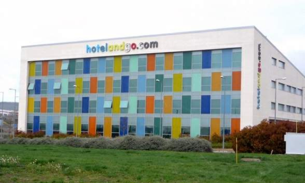 Großes modernes Hotelgebäude mit zahlreichen Fenstern, umgeben von Grünflächen, einem Zaun, Straßenlaternen, geparkten Fahrzeugen, fernen Hügeln und einem bewölkten Himmel; das Wort 'hotel und go' ist auf dem Gebäude zu sehen.