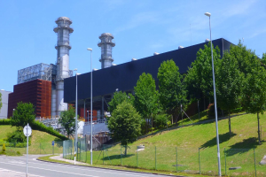 Ein großes Gebäude mit zahlreichen Fenstern, als Kraftwerk identifiziert durch Beschriftung, umgeben von Straßeninfrastruktur, Fahrzeugen, Vegetation und einem bewölkten Himmel.