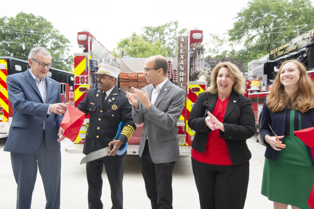 Gruppe von Menschen bei einer Eröffnungszeremonie der Alexandria Fire Department, die vor einem Feuerwehrauto klatschen und lächeln, während zwei Personen eine Schere und ein rotes Band halten.