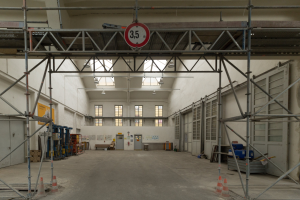 Großer Lagerraum mit ausgedehnten Gerüsten, Verkehrskegeln, Metallstangen, einem Schild, Maschinen, einer Tür, Fenstern, Wänden, Deckenlampen und Dach.