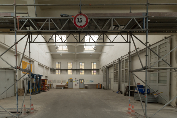 Großer Lagerraum mit ausgedehnten Gerüsten, Verkehrskegeln, Metallstangen, einem Schild, Maschinen, einer Tür, Fenstern, Wänden, Deckenlampen und Dach.