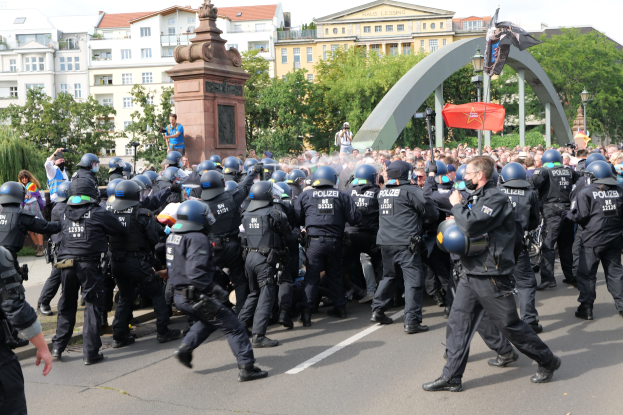 Polizeibeamte in Einsatzausrüstung gehen eine Straße entlang, umgeben von einer Menge, mit Bäumen, Gebäuden, Laternenmasten, einem Bogen, einer Statue und einer Flagge im Hintergrund unter einem bewölkten Himmel.