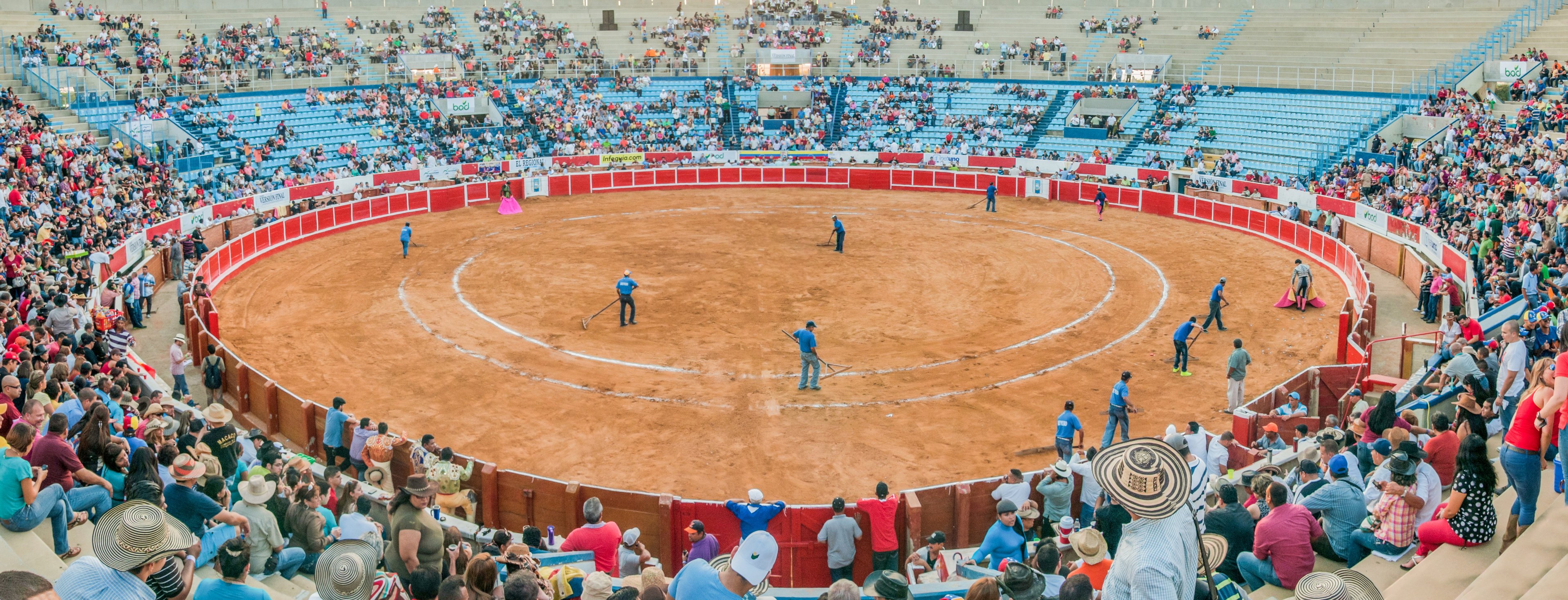 Eine große Menschenmenge schaut bei einem Stierkampf im Stadion zu, wobei einige stehen, sitzen oder Gegenstände halten und eine Person unten einen Hut trägt.