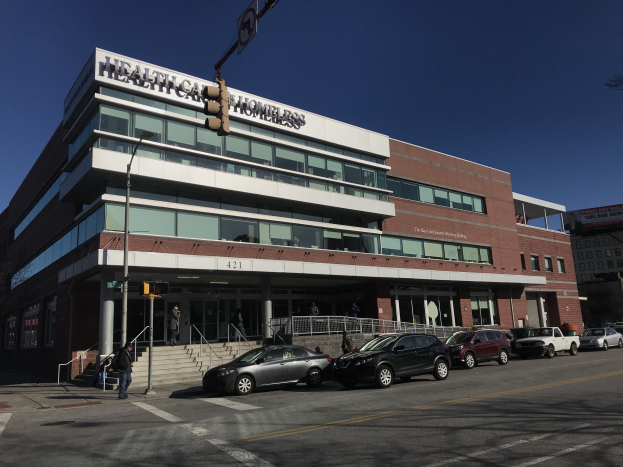 Großes Gebäude mit der Aufschrift 'New York City Health Care Homeless Center' mit parkenden Autos davor, umgeben von Straßeninfrastruktur und Passanten unter einem klaren blauen Himmel.