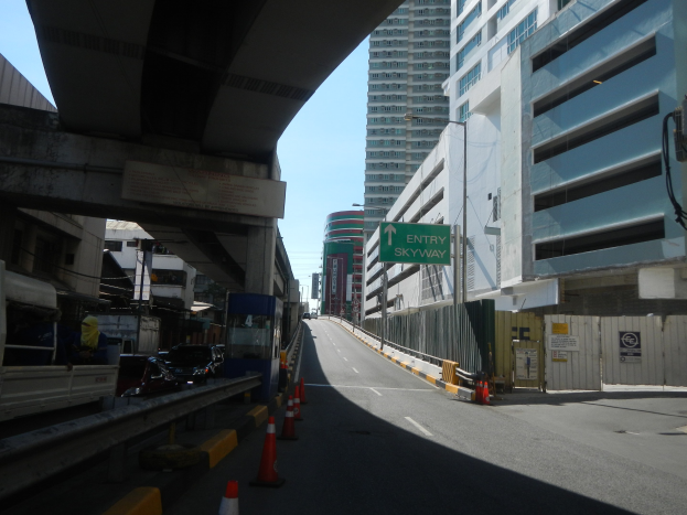 Stadtstraße mit Verkehrskegeln, Fahrzeugen, einer Brücke auf der linken Seite, Gebäuden mit Namensschildern, Laternenpfählen und einem Himmel als Hintergrund.