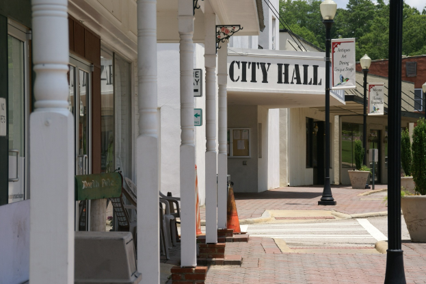 Rathaus mit Gebäuden, Straßenmasten, Straßenlaternen, Namenschildern, Kraftfahrzeugen, Verkehrskegeln, Stühlen, Topfpflanzen, Bäumen und einem bewölkten Himmel im Hintergrund.