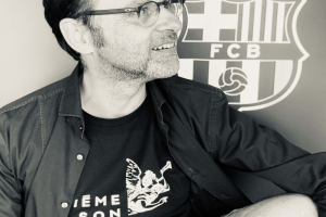 Ein Mann mit Brille und schwarzem Hemd sitzt vor einem schwarz-weißen FC Barcelona-Logo und sieht glücklich aus.