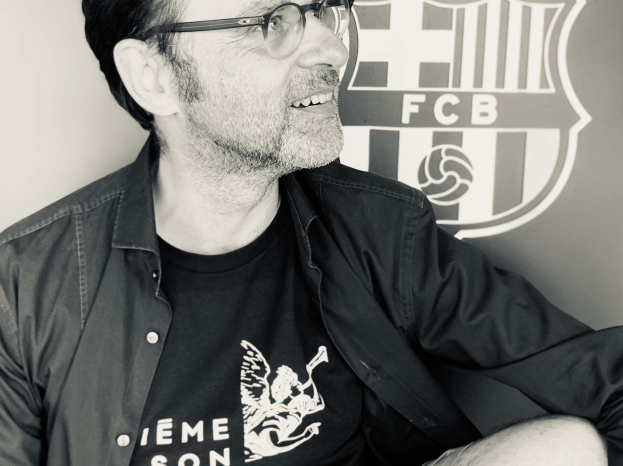 Ein Mann mit Brille und schwarzem Hemd sitzt vor einem schwarz-weißen FC Barcelona-Logo und sieht glücklich aus.